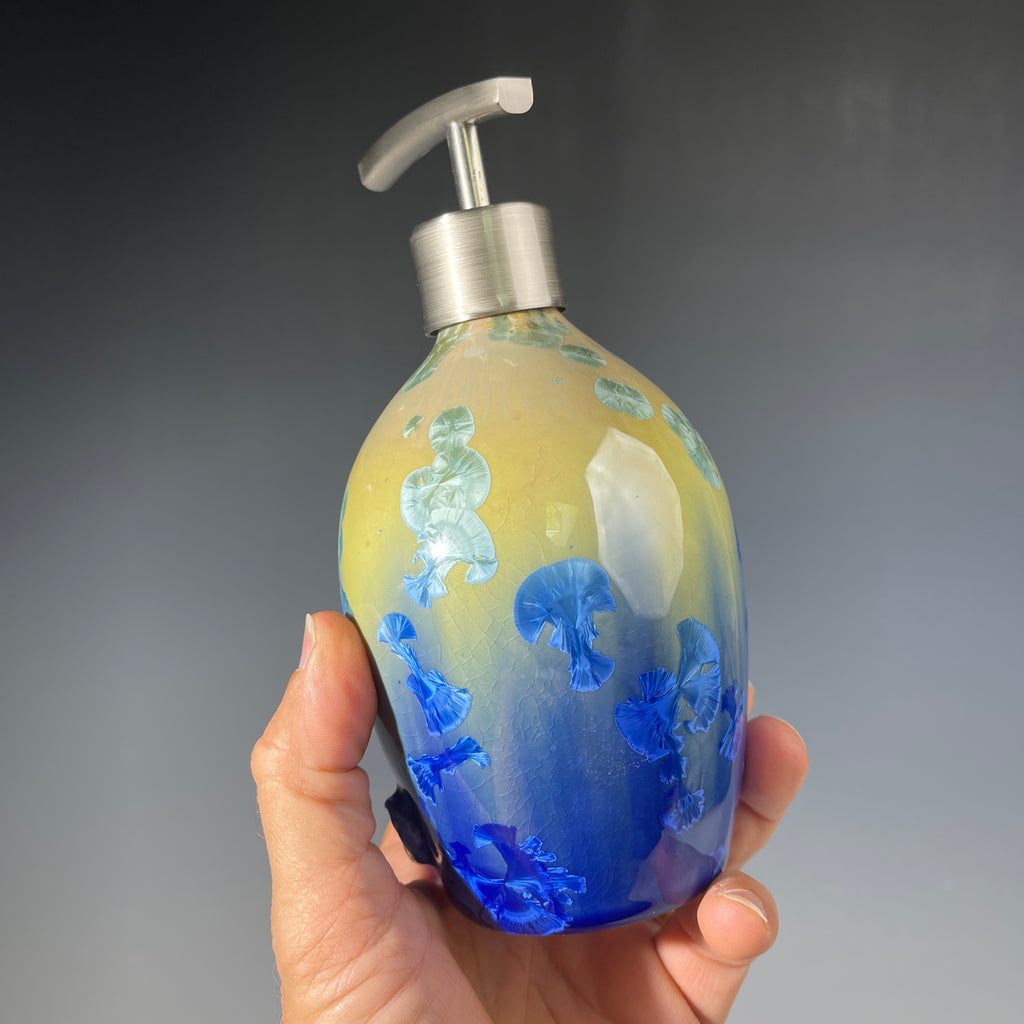 A hand holding a chartreuse to cobalt crystalline glazed soap dispenser against a dark grey background