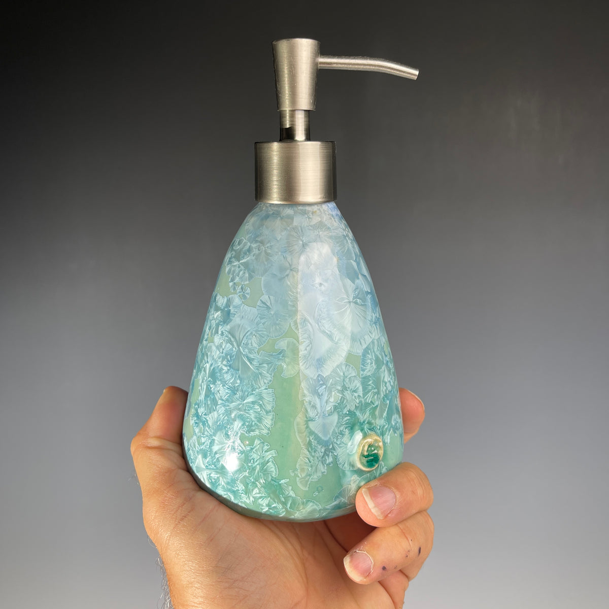 A functional and decorative soap dispenser with a crystalline ceramic  body and a brushed stainless steel pump, shown on a neutral gray background.