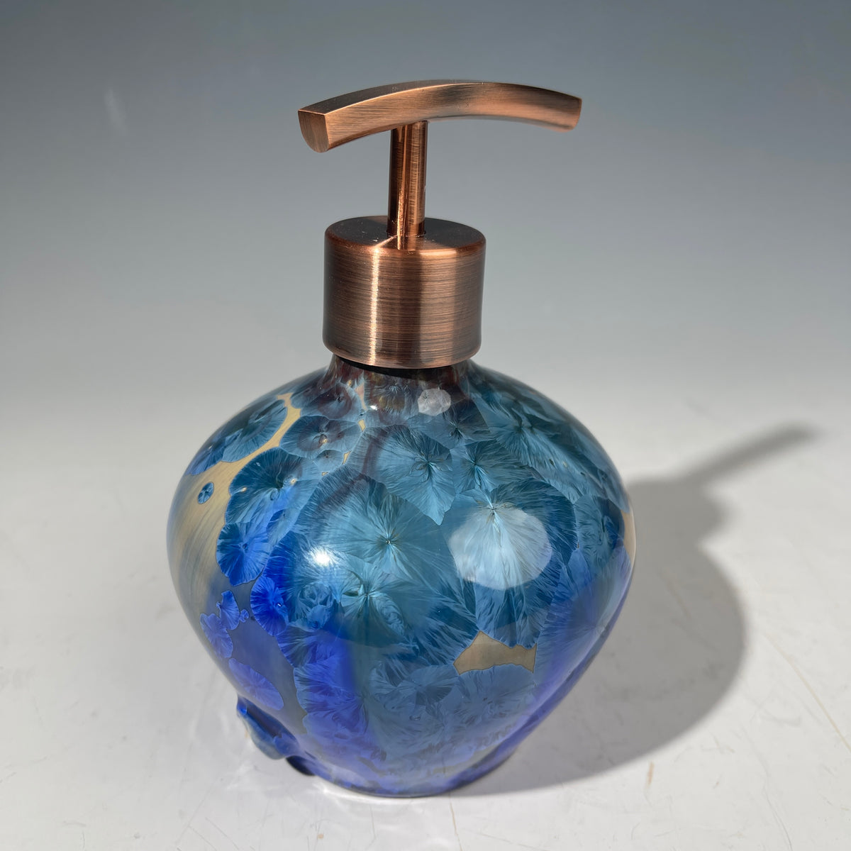 A decorative handmade ceramic functional soap dispenser bottle with a glossy blue and gold crystalline pattern, featuring a copper-finished pump dispenser