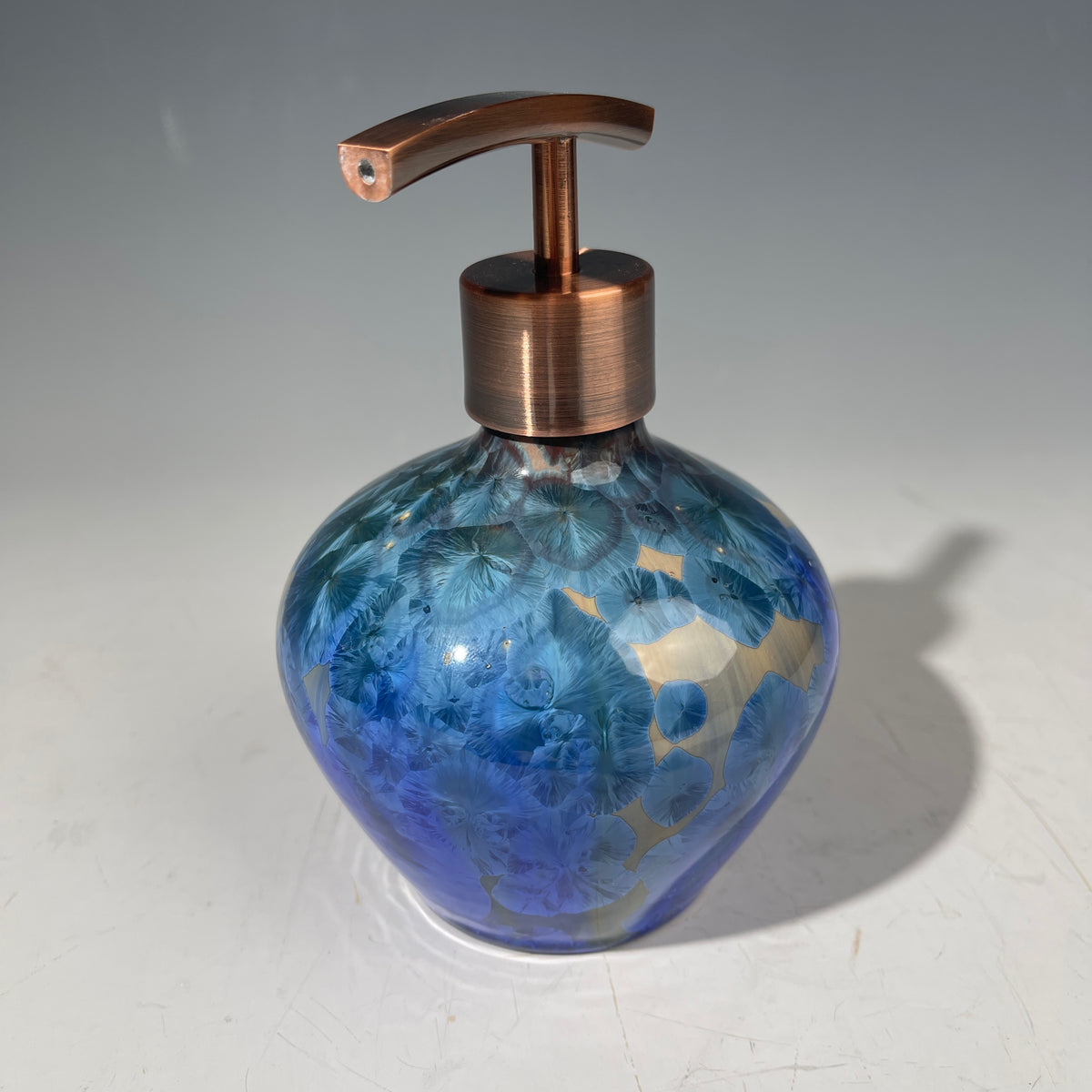 A decorative handmade ceramic functional soap dispenser bottle with a glossy blue and gold crystalline pattern, featuring a copper-finished pump dispenser