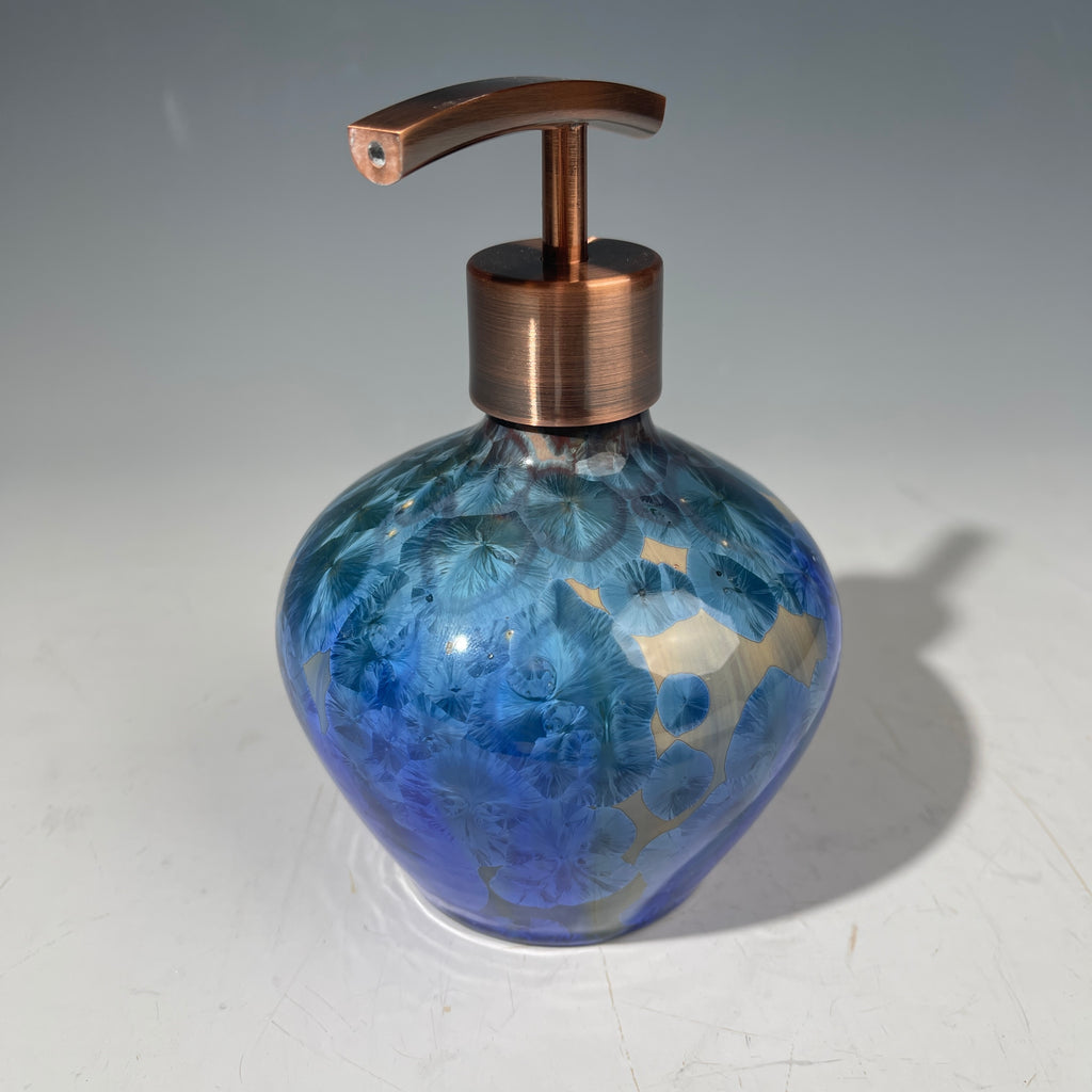 A decorative handmade ceramic functional soap dispenser bottle with a glossy blue and gold crystalline pattern, featuring a copper-finished pump dispenser