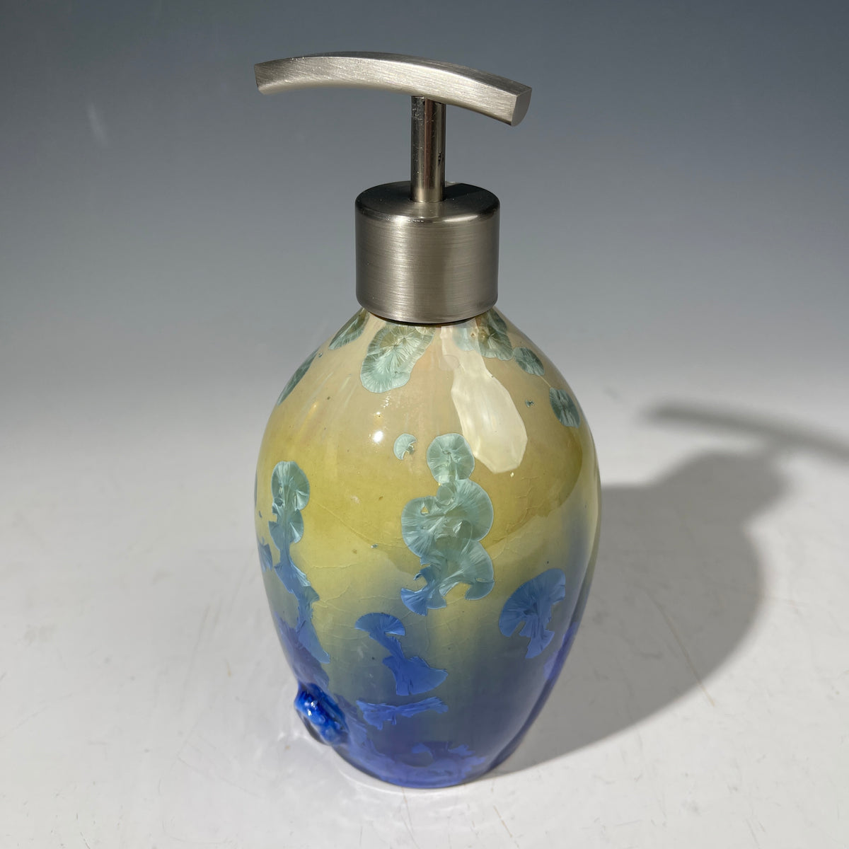 A chartreuse to cobalt crystalline glazed soap dispenser by the artist Hong Rubinstein against a gradient gray background