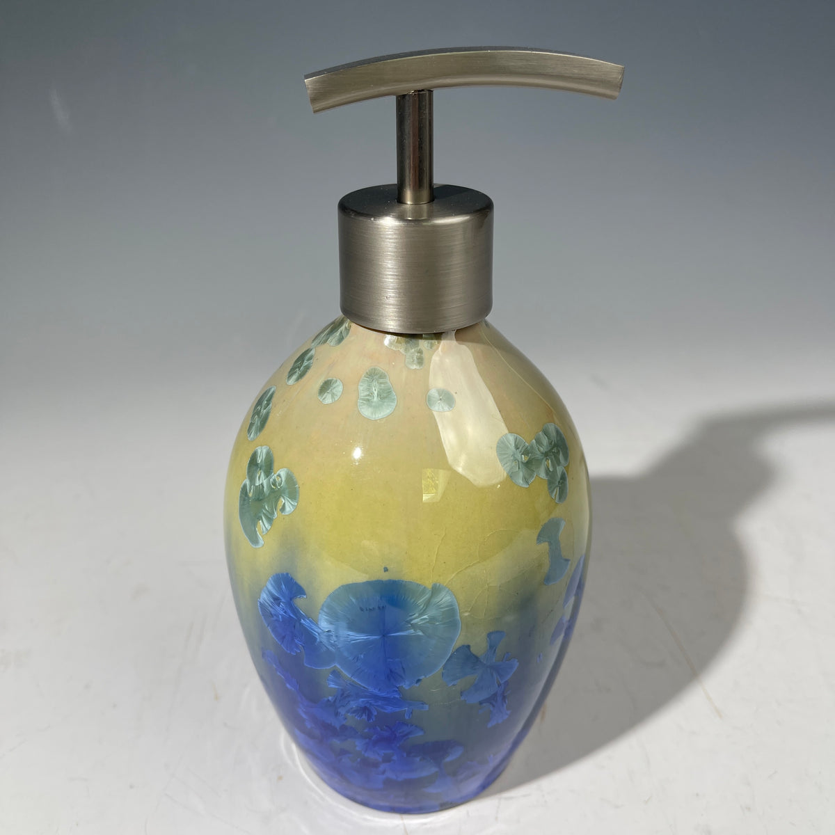 A chartreuse to cobalt crystalline glazed soap dispenser by the artist Hong Rubinstein against a gradient gray background