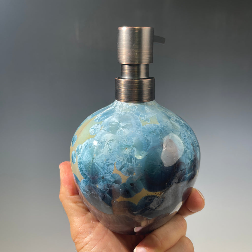 A close-up of a decorative ceramic soap pump featuring a light blue teal and maroon charcoal crystalline glaze