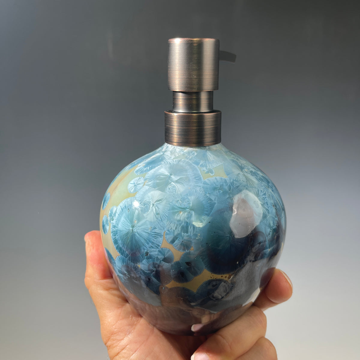 A close-up of a decorative ceramic soap pump featuring a light blue teal and maroon charcoal crystalline glaze