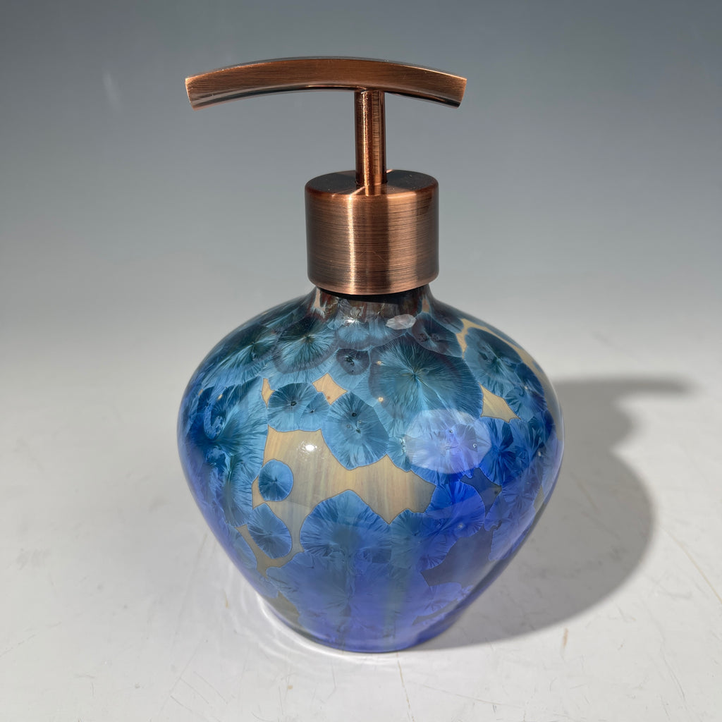 A decorative handmade ceramic functional soap dispenser bottle with a glossy blue and gold crystalline pattern, featuring a copper-finished pump dispenser