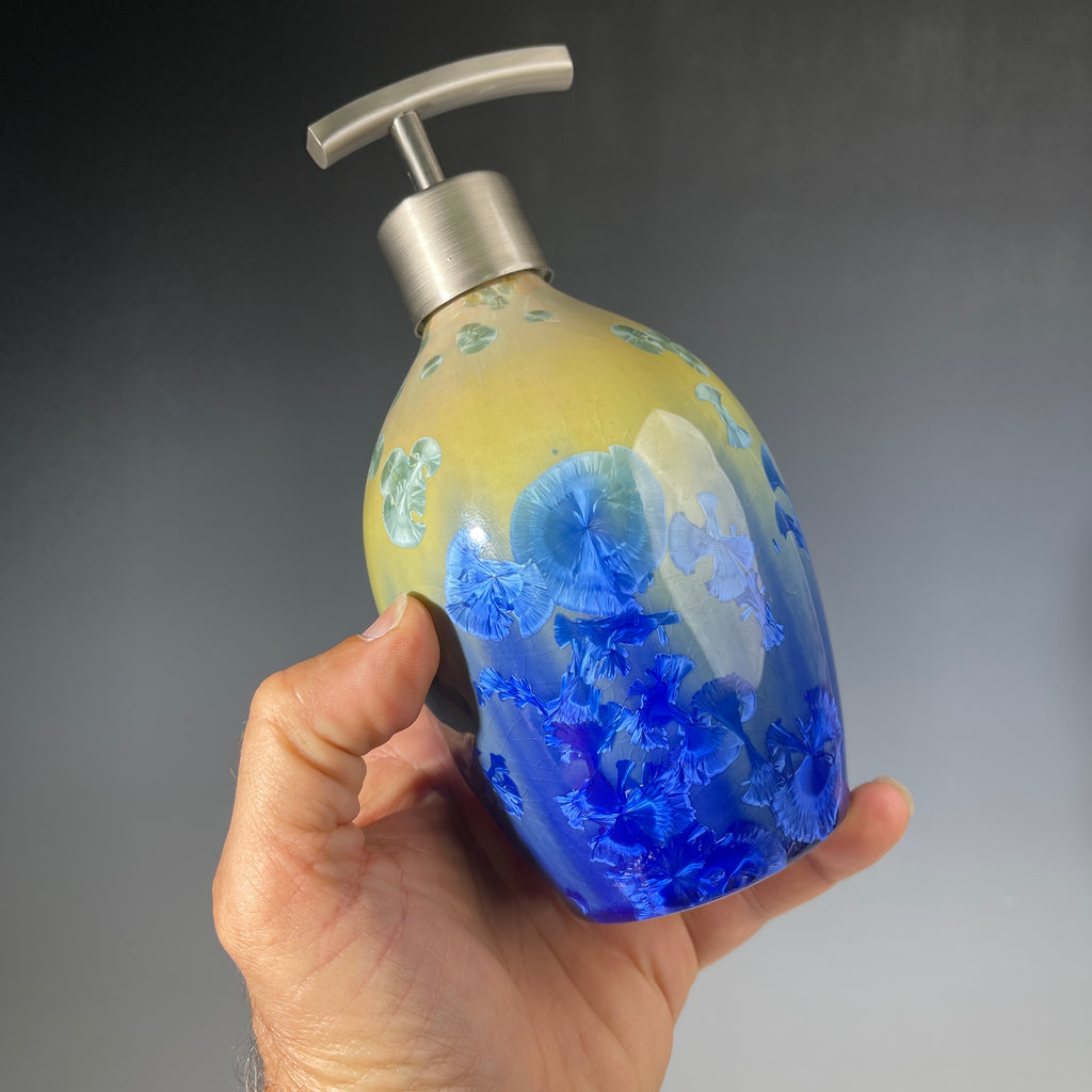 A hand holding a chartreuse to cobalt crystalline glazed soap dispenser against a dark grey background