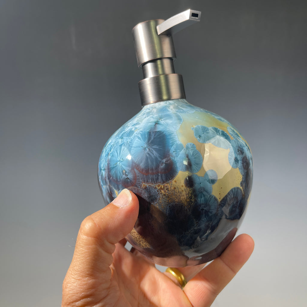 A hand holding a round soap dispenser bottle with a blue, gold, and black crystalline pattern, featuring a brushed silver metal pump dispenser against a gray gradient background.