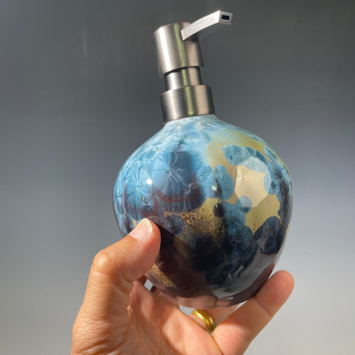 A hand holding a round soap dispenser bottle with a blue, gold, and black crystalline pattern, featuring a brushed silver metal pump dispenser against a gray gradient background.