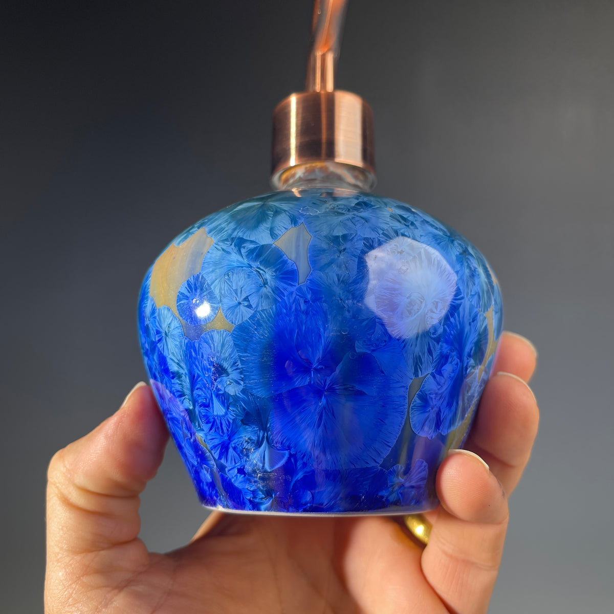 A close up of a  decorative handmade ceramic functional soap dispenser bottle with a glossy blue and gold crystalline pattern, featuring a copper-finished pump dispenser