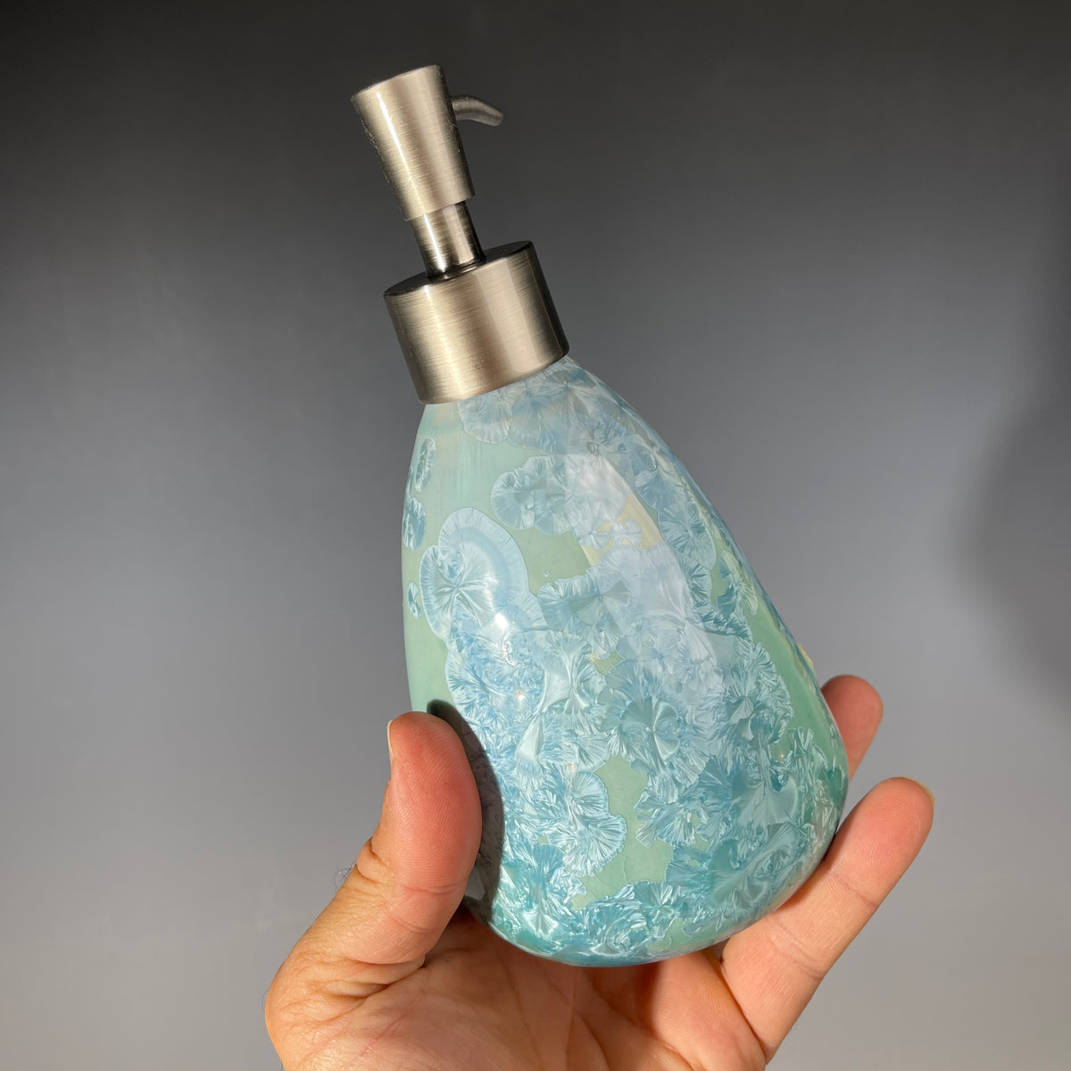 A functional and decorative soap dispenser with a crystalline ceramic  body and a brushed stainless steel pump, shown on a neutral gray background.