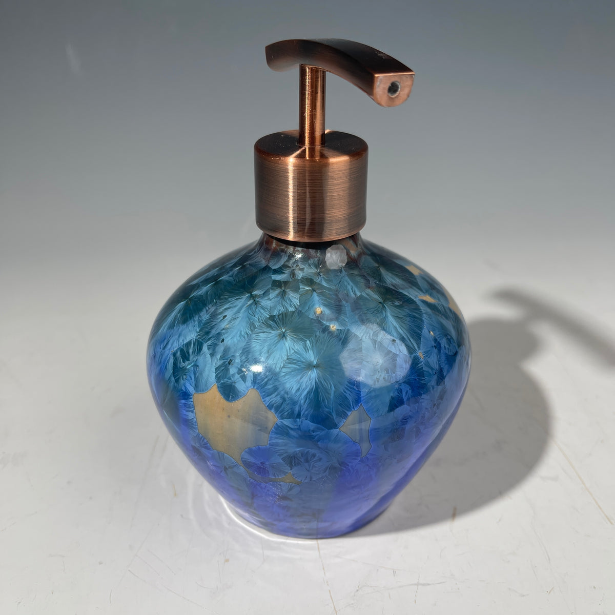 A decorative handmade ceramic functional soap dispenser bottle with a glossy blue and gold crystalline pattern, featuring a copper-finished pump dispenser
