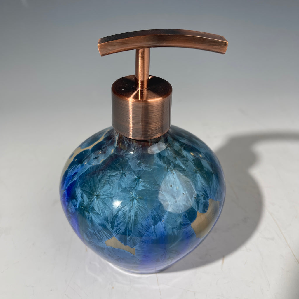 A decorative handmade ceramic functional soap dispenser bottle with a glossy blue and gold crystalline pattern, featuring a copper-finished pump dispenser