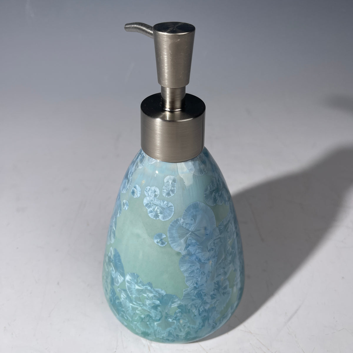 A functional and decorative soap dispenser with a crystalline ceramic  body and a brushed stainless steel pump, shown on a neutral gray background.