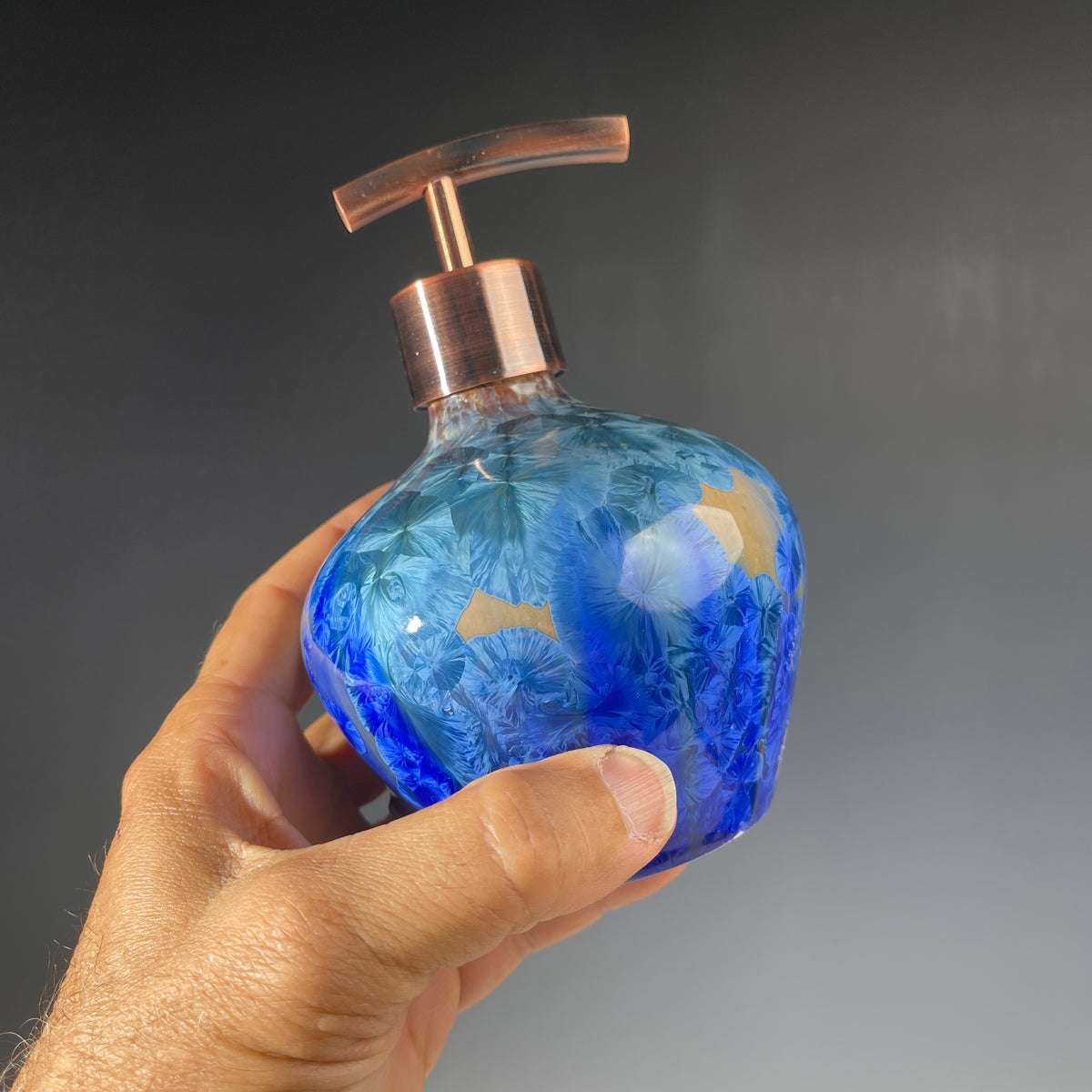 A decorative handmade ceramic functional soap dispenser bottle with a glossy blue and gold crystalline pattern, featuring a copper-finished pump dispenser