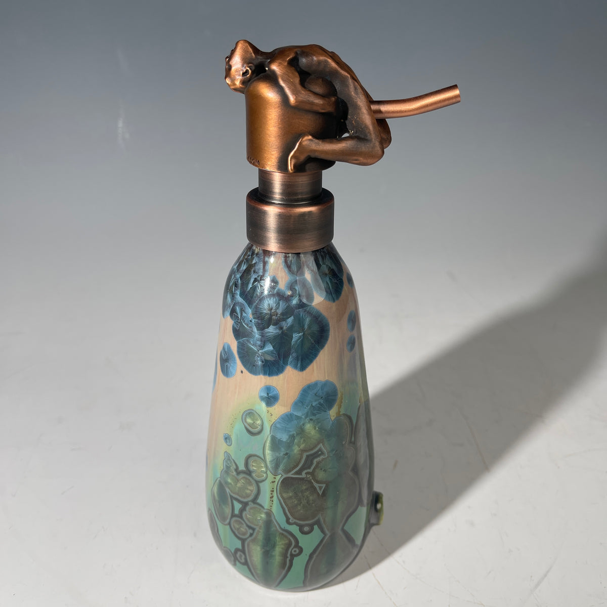 A decorative  and functional ceramic  bottle with a copper-colored metal pump dispenser topped by a sculpted figure, set against a neutral gray background.
