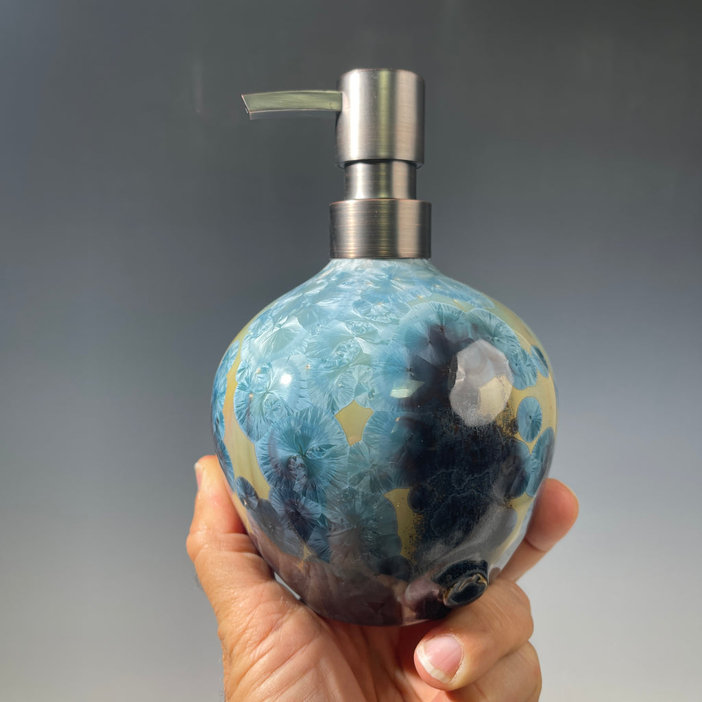 A close-up of a decorative ceramic soap pump featuring a light blue teal and maroon charcoal crystalline glaze