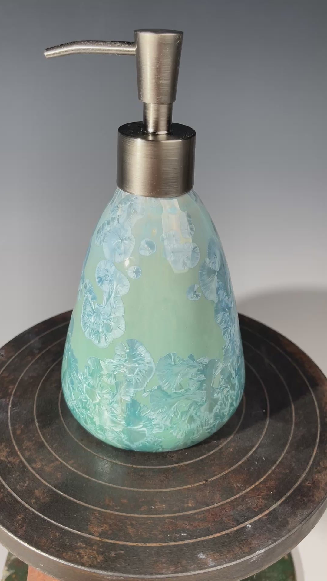 A functional and decorative soap dispenser with a crystalline ceramic  body and a brushed stainless steel pump, shown on a neutral gray background.