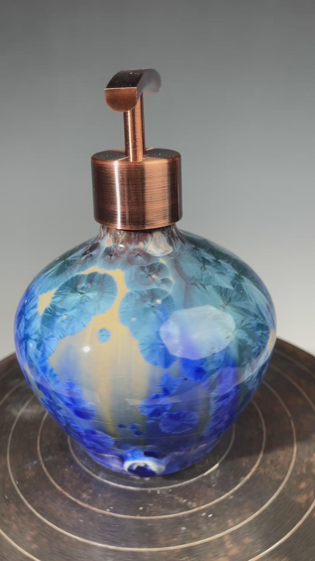 video of a  decorative handmade ceramic functional soap dispenser bottle with a glossy blue and gold crystalline pattern, featuring a copper-finished pump dispenser
